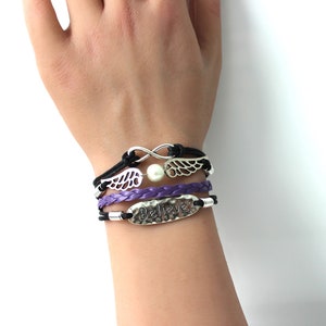May include: A set of three bracelets with a purple braided leather bracelet, a black leather bracelet with a silver infinity symbol and a pearl, and a black leather bracelet with a silver charm that says "Believe".