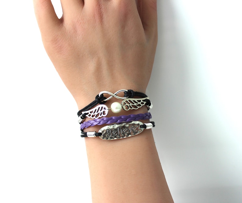 May include: A set of three bracelets with a purple braided leather bracelet, a black leather bracelet with a silver infinity symbol and a pearl, and a black leather bracelet with a silver charm that says "Believe".