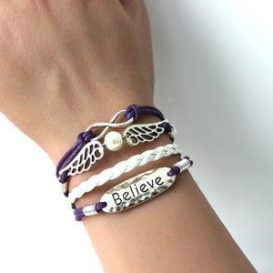 May include: A layered bracelet set with a white braided cord, a purple cord, and silver charms. The charms include a pearl, infinity symbol, and wings with the word "Believe" on a silver tag.