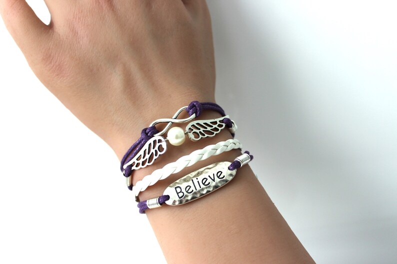 May include: A layered bracelet set with a white braided cord, a purple cord, and silver charms. The charms include a pearl, infinity symbol, and wings with the word "Believe" on a silver tag.