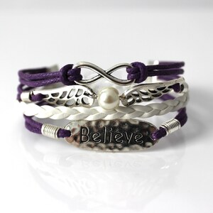 May include: A multi-layered bracelet with purple and white cords, silver infinity symbol, wings, and a pearl charm. The bottom layer features a silver oval charm with the word "Believe" embossed on it.
