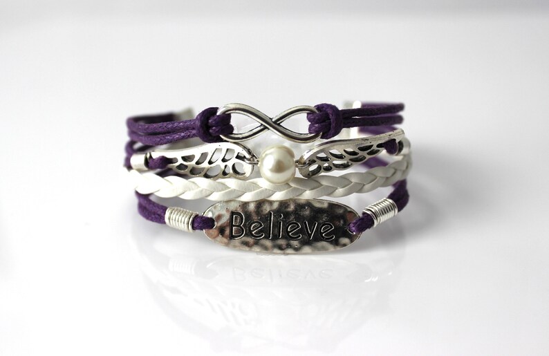 May include: A multi-layered bracelet with purple and white cords, silver infinity symbol, wings, and a pearl charm. The bottom layer features a silver oval charm with the word "Believe" embossed on it.