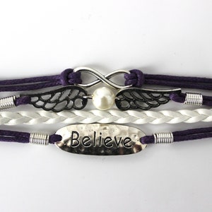 May include: A multi-strand bracelet with purple and white cords, featuring a silver infinity symbol with a pearl, silver wings, and a silver charm with the word "Believe" engraved on it.