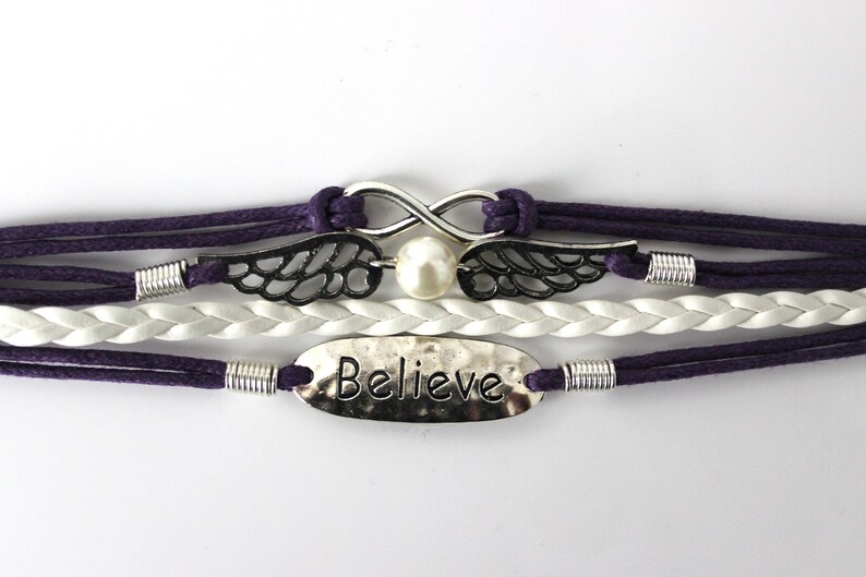 May include: A multi-strand bracelet with purple and white cords, featuring a silver infinity symbol with a pearl, silver wings, and a silver charm with the word "Believe" engraved on it.
