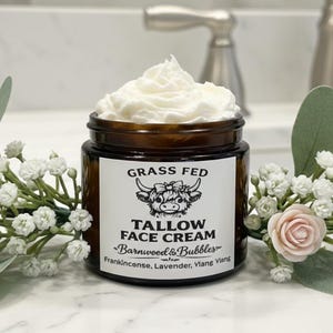 May include: A jar of Grass Fed Tallow Face Cream by Barnwood & Bubbles, with a white label and a cow illustration. The jar is surrounded by white flowers and pink roses. The cream is white and fluffy.