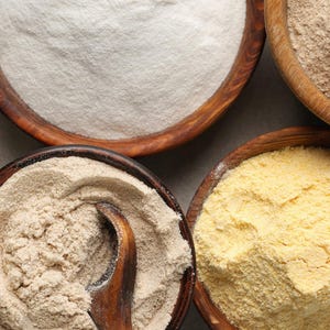 May include: Overhead shot of five wooden bowls filled with different types of flour. The flours vary in color, from bright yellow to off-white and light brown. A wooden spoon is visible in one bowl.