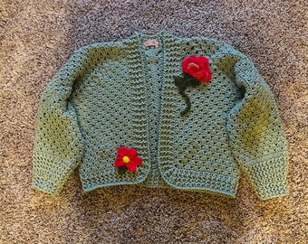 Pode incluir: Um casaco de tricô verde claro com duas flores vermelhas como enfeite. O casaco tem fecho de botão e mangas compridas.