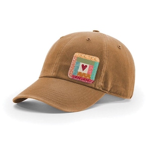 May include: A solid brown baseball cap featuring a square patch on the front panel. The patch displays a colorful patchwork quilt design with various floral and geometric patterns, centered with a small, dark red heart icon on a white background.