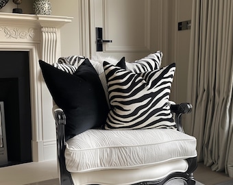 Peut inclure: Un fauteuil blanc avec des pieds et des accoudoirs en bois noir. Le fauteuil est rembourré d'un tissu blanc à motif texturé. Il y a trois coussins sur le fauteuil, deux coussins en velours noir et un coussin à imprimé zèbre.