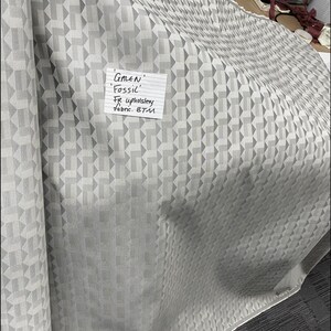May include: A close-up of a silver-grey fabric with a geometric pattern. The fabric is labelled "GALEN Fossil' FIR upholstery Fabric. BTM". The photo was taken under bulb lighting.