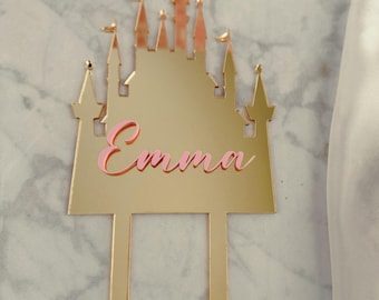 May include: A gold-colored castle cake topper with the name "Emma" in pink script. The castle design features multiple turrets with small flags. The topper has two vertical posts for insertion into a cake.