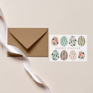 Puede incluir: Una tarjeta blanca con un dise&ntilde;o floral en ocho huevos y el texto "Feliz Pascua". La tarjeta est&aacute; junto a un sobre marr&oacute;n y una cinta blanca.