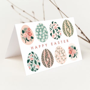 Puede incluir: Una tarjeta blanca con un dise&ntilde;o floral. La tarjeta tiene un patr&oacute;n floral rosa, verde y blanco en los huevos. El texto "Happy Easter" est&aacute; impreso en rosa en la tarjeta.