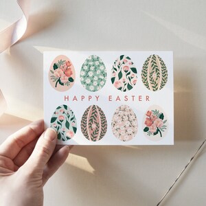 Puede incluir: Una tarjeta blanca con el texto "Happy Easter" en letras rosas. La tarjeta est&aacute; decorada con ocho huevos de color pastel, cada uno con un patr&oacute;n floral diferente.