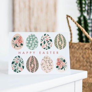Puede incluir: Una tarjeta blanca con el texto "Happy Easter" impreso en rosa. La tarjeta presenta ocho huevos de Pascua ilustrados con motivos florales en tonos rosa, verde y blanco.