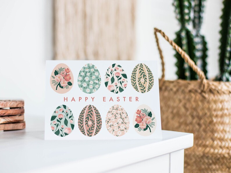 Puede incluir: Una tarjeta blanca con el texto "Happy Easter" impreso en rosa. La tarjeta presenta ocho huevos de Pascua ilustrados con motivos florales en tonos rosa, verde y blanco.