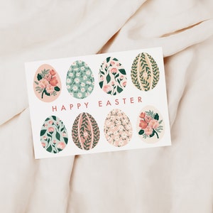 Puede incluir: Una tarjeta blanca con un dise&ntilde;o floral. La tarjeta presenta ocho huevos de Pascua de color rosa pastel y verde con patrones florales. El texto "Feliz Pascua" est&aacute; impreso en rosa en la tarjeta.