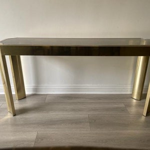 May include: A gold-toned console table with a glass top. The table has a simple, modern design with a rectangular shape and straight legs.