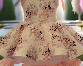 May include: A light pink and white dress with a Disney-themed print featuring Mickey and Minnie Mouse. The dress has a ruffled skirt and straps with bows. The background shows a castle and pink trees, creating a whimsical aesthetic.