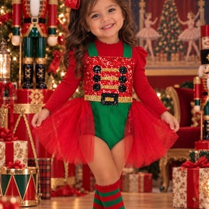 May include: A red long-sleeved shirt worn under a green and red sequined nutcracker-themed romper with a tulle skirt, paired with red and green striped knee-high socks and a red sequined hair bow, featuring black buttons and a gold belt detail.