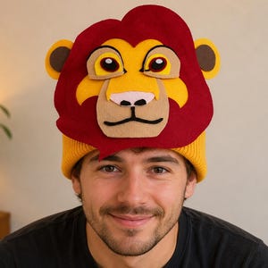 May include: A yellow knit beanie with a lion face design. The lion's mane is red, with yellow and tan accents for the face, eyes, and nose. The beanie is worn by a person wearing a black shirt.