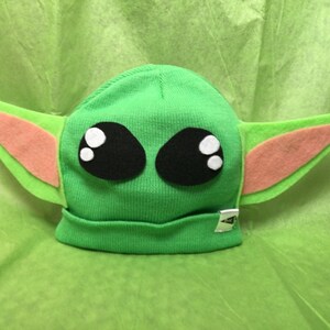 May include: A green knit beanie hat with large black eyes, white highlights, and attached felt ears in green and peach. The hat has a small silver tag on the brim. The hat is designed to resemble a popular character.