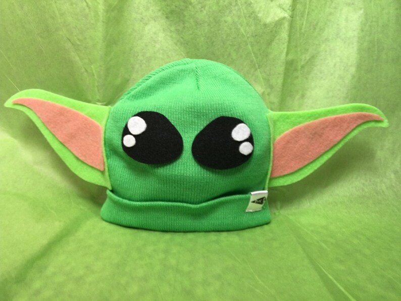 May include: A green knit beanie hat with large black eyes, white highlights, and attached felt ears in green and peach. The hat has a small silver tag on the brim. The hat is designed to resemble a popular character.
