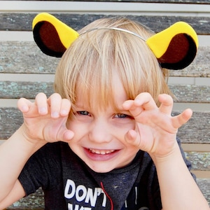 May include: A child wearing a headband with yellow and brown lion ears, and a dark gray t-shirt with the text "DON'T". The child is smiling and making a claw gesture with their hands.
