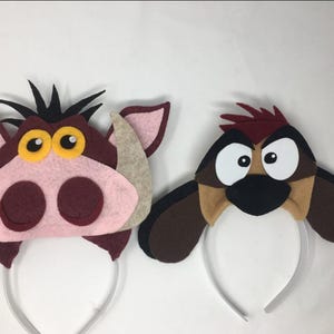 May include: Two character headbands. One is a pink and brown warthog with yellow eyes and black spikes. The other is a brown and tan bird with large white eyes. Both are attached to white headbands.