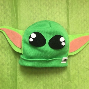 May include: A green knit beanie hat with large black eyes and white highlights, and large green and pink felt ears. The hat is designed to resemble a popular character from a science fiction franchise. The hat is on a green background.