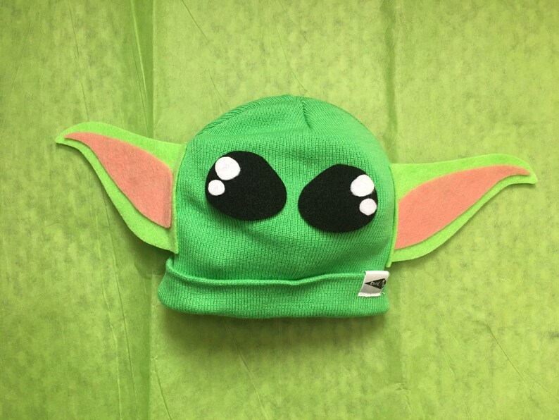 May include: A green knit beanie hat with large black eyes and white highlights, and large green and pink felt ears. The hat is designed to resemble a popular character from a science fiction franchise. The hat is on a green background.