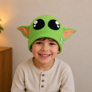 May include: A child wearing a green fleece hat with large black eyes, tan inner ears, and pointed ears. The hat resembles a character from a popular science fiction franchise. The child is smiling and wearing a light-colored long-sleeved shirt.