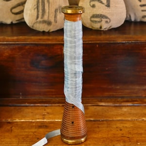 May include: A vintage wooden spool with a brass top and bottom, wrapped with light blue ribbon. The spool is set on a wooden surface, with a wooden box and burlap sacks in the background.