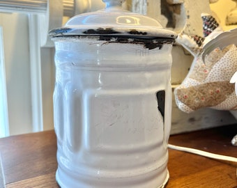 May include: White enamelware canister with a lid, featuring a black rim and a slightly chipped surface. The canister has a ribbed design and is slightly worn.