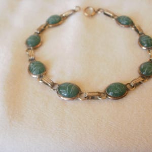 May include: A metal link bracelet featuring eight oval-shaped green stone cabochons, each engraved with a scarab beetle design. The bracelet is constructed with gold-toned metal connectors between each stone and includes a circular clasp.