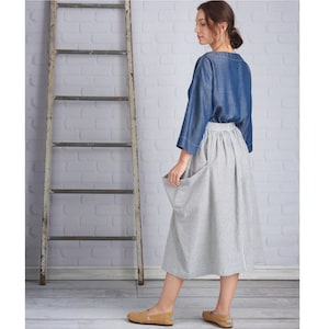 May include: A denim blue top with wide sleeves and a white and gray striped skirt with large pockets. The outfit is paired with tan slip-on shoes. A wooden ladder leans against a white brick wall.