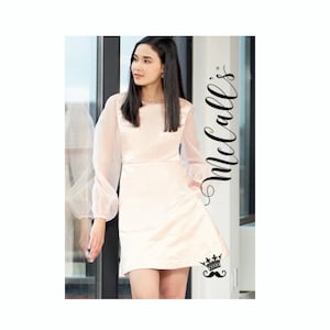 Op de afbeelding: Een lichtperzikkleurige jurk met lange, doorschijnende mouwen. De jurk heeft een vierkante halslijn en een getailleerd lijfje dat iets uitloopt bij de taille. De tekst "McCall's" staat verticaal aan de rechterkant van de afbeelding.