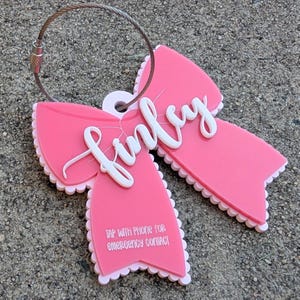 May include: A pink luggage tag in the shape of a bow with the name "Finley" in white script. The tag has a scalloped edge and includes the text "TAP WITH PHONE FOR EMERGENCY CONTACT". A metal loop is attached for securing the tag.