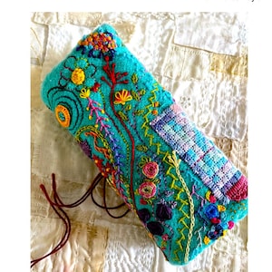 Op de afbeelding: Een turquoise naaldenboek met kleurrijke borduurwerk van bloemen en wijnstokken. Het boek is vastgebonden met een bruin koord en ligt op een witte stoffen achtergrond. De tekst "Needlebook Garden" en "Your secret garden of stitching" staat boven de afbeelding. De tekst "With Andrea Taylor" staat onder de afbeelding.