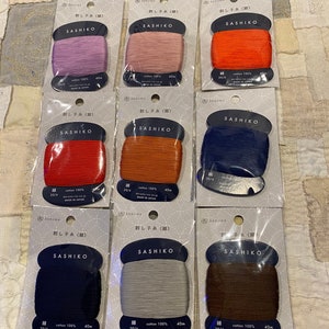 May include: Nine spools of Sashiko thread in various colors, including red, orange, blue, brown, and gray. Each spool is labeled with the brand name "DARUMA" and the text "SASHIKO" in Japanese. The spools are arranged in three rows of three.
