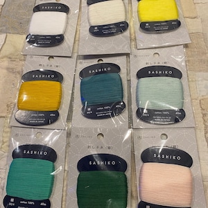 May include: Nine spools of thread for sashiko embroidery. The spools are labelled with the brand name "Daruma" and the text "Sashiko". The thread colours are white, yellow, light green, dark green, and pink.