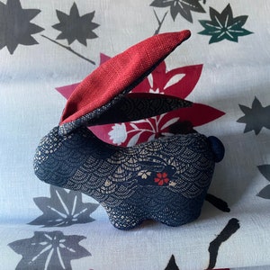 May include: A plush toy rabbit with a red fabric ear and a blue and white patterned body. The rabbit is sitting on a white fabric background with a red and black floral pattern.