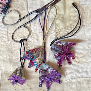 Könnte beinhalten: Drei handgefertigte Stoffblumen-Anhänger mit Perlenakzenten. Die Anhänger sind lila, rosa und blau mit Blumenmustern. Jeder Anhänger ist an einer Schnur und einem Metallring befestigt.