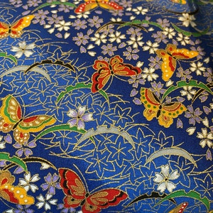 May include: Close-up of a blue fabric featuring a repeating pattern of butterflies, flowers, and leaves. The butterflies have red, yellow, and black wings. The flowers are white and purple, and the leaves are green and gold.