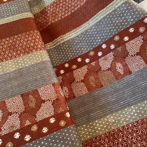 Op de afbeelding: Gerolde stof met een herhalend patroon van geometrische vormen, bloemmotieven en strepen in tinten roest, grijs en beige. De stof lijkt een geweven materiaal te zijn, mogelijk voor quilten of andere knutselprojecten.