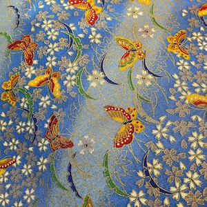 May include: Close-up of a blue fabric featuring a repeating pattern of butterflies and floral motifs. The butterflies are detailed with red, yellow, and gold. White and purple flowers are interspersed with green and gold accents.