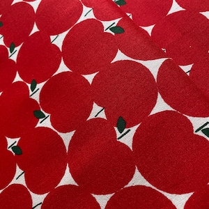 May include: Close-up of red fabric featuring a repeating apple design. Each apple is a bright red with a white triangle and a small green leaf. The fabric has a slight sheen.