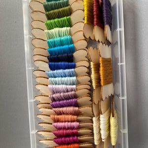 Puede incluir: Un recipiente de plástico transparente lleno de hilos de bordar coloridos. Los hilos están enrollados en carretes de madera en una variedad de colores, incluyendo verde, azul, morado, naranja y rojo. Los carretes están dispuestos en filas.