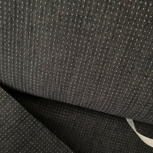May include: Close-up of dark grey fabric with a repeating pattern of small white dots. The fabric is folded, revealing its texture and the subtle contrast between the dark base and the lighter dots. The material appears to be a woven textile.