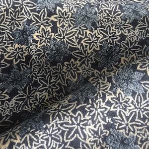 May include: Close-up of a fabric with a repeating floral pattern. The design features cream-coloured, stylised leaves against a dark navy blue background. The fabric has a textured appearance, with areas of lighter blue and white speckling.